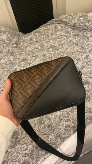 Fendi axelväska med FF-mönster - Snygg axelväska från Fendi med klassiskt FF-logomönster i brunt och mörkbrunt. Väskan har en rektangulär form, svart justerbar axelrem med Fendi-tryck och silverfärgad dragkedja. Materialet är skinn och canvas, perfekt för dig som gillar lyxiga detaljer och streetstyle.