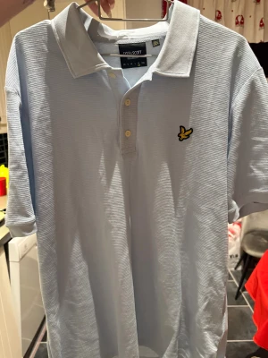 Ljusblå pikétröja från Lyle & Scott - Snygg ljusblå pikétröja från Lyle & Scott i relaxed fit. Tröjan har klassisk krage, två knappar och det ikoniska gula fågelbroderiet på bröstet. Materialet är bomull med en diskret strukturerad yta. Perfekt för dig som gillar stilrena och bekväma plagg.