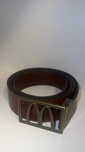 Brunt McDonald's skärp i skinn - Säljer ett brunt bälte i skinn med en unik metallspänne som har den klassiska McDonald's-loggan. Bältet har en stilren och enkel design med rektangulärt spänne i antik mässing-look. Perfekt för dig som vill sticka ut med en ikonisk detalj.