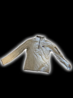 Grå fleece half zip från Patagonia - Snygg grå fleece-tröja från Patagonia med half zip och lång ärm. Tröjan har en liten ficka med dragkedja på ena ärmen och ett diskret logomärke på bröstet. Perfekt för kyliga dagar och outdoor-häng.