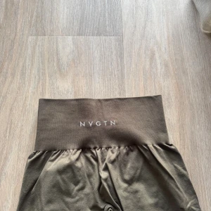 Gröna leggings från NVGTN - Snygga gröna leggings från NVGTN med bred, hög midja och diskret logga framtill. Stretchigt och mjukt material som sitter skönt och formar sig efter kroppen. Perfekta för gymmet eller chill hemma. Färgen är neutral och lätt att matcha med andra plagg.