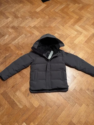 Canada Goose macmillan - Säljer en grå pufferjacka från Canada Goose med huva och snyggt svart märke på ärmen. Jackan har dragkedja och knappar framtill, två sidofickor och quiltad design. Perfekt för dig som vill ha en stilren och varm jacka med street vibe.