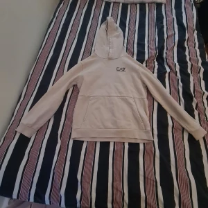 Beige hoodie från EA7 Emporio Armani - Snygg beige hoodie från EA7 Emporio Armani med huva och stor magficka. Tröjan har lång ärm och en stilren logga på bröstet. Tillverkad i mjuk bomull och polyesterblandning, perfekt för chill dagar. Passar dig som gillar enkel och clean stil.           Vet inte ifall fläcken går bort.