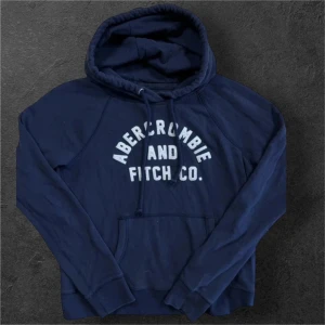 Blå hoodie från Abercrombie & Fitch - Mörkblå hoodie från Abercrombie & Fitch med vit logga framtill. Tröjan har huva med snörning och en stor magficka. Perfekt för en avslappnad och cool stil. Långärmad och i mjukt bomullsmaterial.