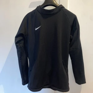 Svart Nike hoodie med huva - Svart hoodie från Nike med klassisk vit swoosh på bröstet. Tröjan har huva, långa ärmar och praktiska fickor framtill. Perfekt för träning eller chill, enkel och stilren design i mjukt material.