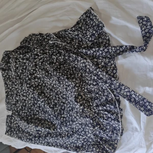 Blommig omlottkjol från SHEIN - Supersöt svart omlottkjol från SHEIN med vitt blommönster och volangkant. Kjolen har knytband i midjan och dragkedja i sidan för enkel påklädning. Perfekt för dig som gillar en feminin och trendig stil.