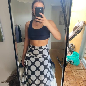 Svartvit prickig omlottkjol - Säljer en svartvit omlottkjol med stora vita prickar. Kjolen har volangkant nertill och knyts i sidan för en snygg passform. Längden är mitt på vaden och tyget är lätt och flowy, perfekt för varmare dagar.