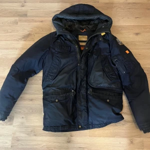 Mörkblå Parajumpers vinterjacka - Säljer en mörkblå Parajumpers vinterjacka med huva och pälsfoder. Jackan har flera stora fickor, coola detaljer som patch på ärmen och gul rem vid kragen. Perfekt för kalla dagar och riktigt snygg streetstil.