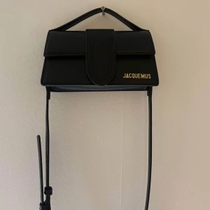 Axelväska - Säljer en svart axelväska i stilren design med guldigt logotyptryck framtill. Väskan har ett avlångt, rektangulärt format, smal axelrem och ett lock med bred detalj. Perfekt för dig som gillar minimalistisk och trendig look. HELT NY