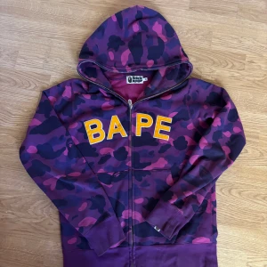 BAPE lila camo hoodie med huva - Säljer en fet hoodie från BAPE i lila och mörklila camouflage med stora gula BAPE-bokstäver på bröstet. Tröjan har huva, dragkedja och ribbade muddar. Perfekt för dig som vill sticka ut med streetwear. Materialet är mjuk bomull och passformen är normal. Den är äkta har bevis 