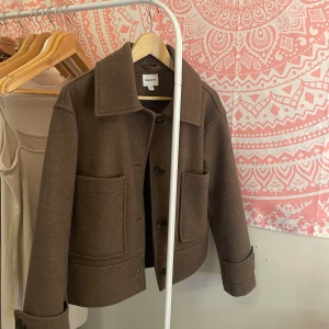 Brun kappa - Snygg brun jacka från Kappahl i ullmix, modell med stora fickor framtill och klassisk krage. Storlek xs. Jackan har knäppning med svarta knappar och är lite boxig i passformen. Perfekt för hösten och vintern! 