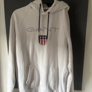 Vit hoodie från GANT med broderad logga - Vit hoodie från GANT med broderad logga och amerikanskt inspirerat emblem på bröstet. Tröjan har huva med snörning, känguruficka och långa ärmar. Tillverkad i mjuk bomull, perfekt för chill dagar. Snygg och klassisk streetstil.Har även någon fläck och sliten vid armarna 