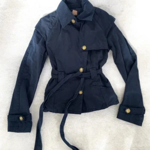 Mörkblå trenchcoat från Patrizia Pepe - Snygg mörkblå trenchcoat från Patrizia Pepe med bälte i midjan och stora guldknappar. Jackan har klassisk krage, långa ärmar och är tillverkad i 100% bomull. Perfekt för dig som vill ha en stilren och tidlös look.