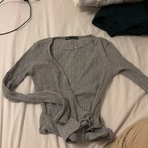 Grå ribbad omlottröja från Zara - Supersnygg grå ribbad omlott tröja från Zara i storlek S. Koftan har långa ärmar, knytband i midjan och en v-ringad omlottfront. Perfekt att slänga på sig på med ett par jeans för en chill look.