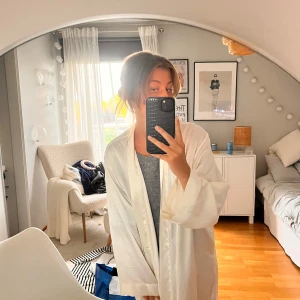 Silkes morgonrock från Victoria's Secret - Säljer en elegant vit omlottklänning från Victoria's Secret i satinliknande material. Klänningen har långa ärmar, knytband i midjan och en mjuk, glansig finish. Perfekt för dig som gillar en lyxig och chill vibe. PS en defekt förekommer som jag tog bild på