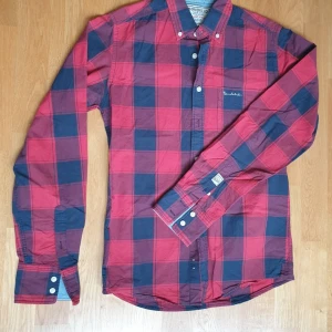 Röd och svart rutig skjorta Bondelid - Snygg rutig skjorta från Bondelid i rött och svart. Skjortan har klassisk krage, långa ärmar och knäppning framtill. Tillverkad i bomull och har en broderad logga på bröstet. Perfekt för en avslappnad men stilren look.