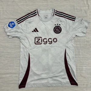 Ajax bortatröja Adidas L senaste säsongen  - Ajax bortatröja i storlek L från Adidas. Tröjan är vit med vinröda detaljer, geometriskt mönster och klassiska Adidas-ränder på axlarna. Ajax klubbmärke och Ziggo-logga på bröstet. Tillverkad i lätt och ventilerande material (Aeroready).