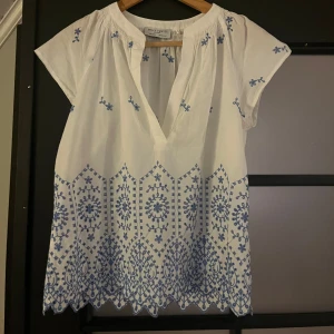 Vit blus med blå broderi Holly & Whyte - Superfin vit blus från Holly & Whyte med blått broderat mönster och v-ringning. Blusen har korta ärmar och dekorativa broderier både upptill och längs fållen. Perfekt för en fräsch och somrig look!