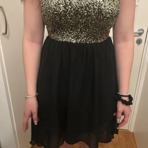 Svart och guldig paljettklänning - Snygg kortklänning med svart, flowy kjol och överdel täckt av guldiga paljetter. Klänningen är från 90-2000 talet. Klänningen har ett litet hål i botten av det svarta men märks knappt och den har ett litet snit. Klänningen är ärmlös och har öppen rygg med dragkedja bak. Perfekt för fest och bal! Materialet känns lätt och glansigt, och paljetterna ger en riktigt glam känsla. 