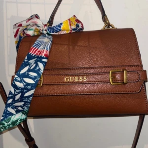 Brun axelväska från Guess  - Snygg brun axelväska från Guess i skinn med guldfärgade detaljer och spännen. Väskan har ett stilrent, fyrkantigt format och en färgglad scarf knuten vid handtaget som extra detalj. Perfekt storlek för det viktigaste och ger en trendig touch till din outfit. Ny kostar 2500kr
