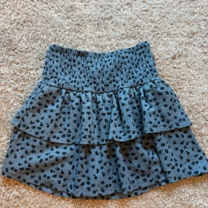 Blommig volangkjol från Zara - Supersöt kort kjol från Zara med volanger och blommigt mönster i svart på blå bakgrund. Kjolen har en smockad midja som sitter skönt och ger en snygg passform. 