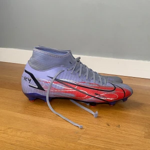 Nike Mercurial fotbollsskor KM lila/röd - Säljer ett par Nike Mercurial fotbollsskor i lila och rött med svarta detaljer och KM-logga. (Kylian  mbappe)  Skorna har snörning, hög krage och är tillverkade i syntetmaterial för bästa grepp på planen. Perfekta för dig som vill sticka ut med stil på fotbollsplanen.