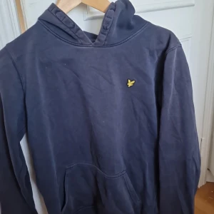Mörkblå hoodie från Lyle & Scott - Säljer en mörkblå hoodie från Lyle & Scott med klassisk gul logga på bröstet. Tröjan har huva med snörning och en stor magficka framtill. Perfekt för dig som gillar en enkel och stilren look. lite trasig på handleden fast syns inte. Pris kan diskuteras!!