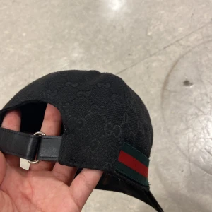 Svart Gucci keps med GG-mönster - Snygg svart keps från Gucci med diskret GG-monogram över hela tyget och klassiska gröna och röda ränder på sidan. Justerbar rem bak i skinn och mjukt bomullsmaterial. Perfekt för dig som gillar exklusiva accessoarer med streetkänsla.