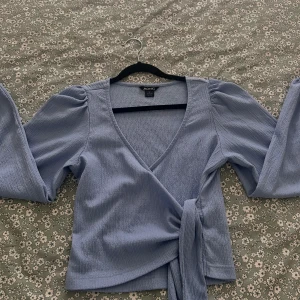 Ljusblå omlottopp från Monki - Supersöt ljusblå omlottopp från Monki med långa puffärmar och breda knytband i midjan. Toppen har v-ringning och är gjord i ett strukturerat, mjukt tyg som känns riktigt skönt mot huden. Perfekt för dig som gillar trendiga och feminina detaljer.