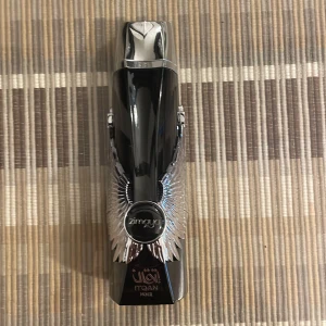 Zimaya Itqan Noir Eau de Parfum - Snygg parfymflaska på 100 ml från Zimaya, Itqan Noir. Flaskan är svart med silvriga vingar på sidorna och har en futuristisk, elegant design med metallisk topp. Kommer i en svart kartong med vingmotiv. Perfekt för dig som gillar unika och stilrena detaljer. Använd några gånger ungefär 95-97% kvar.