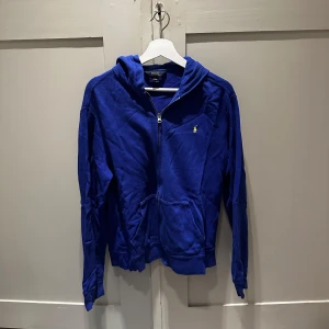Blå hoodie från Polo Ralph Lauren - Blå hoodie från Polo Ralph Lauren med dragkedja och huva. Klassisk gul broderad logga på bröstet och känguruficka framtill. Står XL men skulle säga M