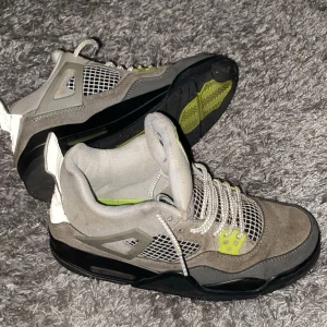 Nike Air Jordan 4 Retro Neon - Nike Air Jordan 4 Retro Neon med grå mocka, svarta detaljer och inslag av neongult. Skorna har vit snörning, meshpaneler och en ikonisk Jumpman-logga på plösen. Snygg kontrast mellan olika grå nyanser och neongula accenter. Perfekta för dig som gillar streetstyle.priset kan diskuteras