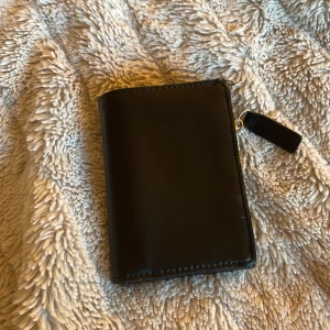 Svart plånbok i skinn från Åhléns - En stilren svart plånbok i skinn från Åhléns med flera kortfack, myntfack med dragkedja och plats för sedlar. Klassisk design med tryckknappsstängning och smidig storlek som passar perfekt i fickan eller väskan.