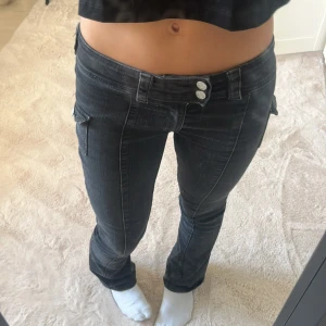 Svarta bootcut jeans med låg midja - Säljer ett par svarta jeans med låg midja och raka ben. Byxorna har coola cargofickor på sidorna med knappdetaljer och dubbla knappar i midjan. Jeansen är i stretchigt denim och har en snygg, mörk tvätt som passar till allt. På bilden 164 cm lång💕