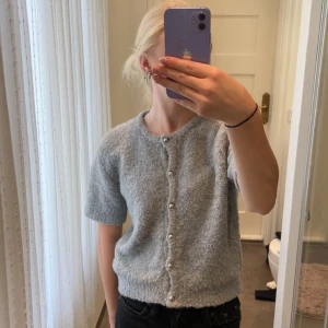 Ljusgrå kofta från Gina Tricot - Supermjuk ljusgrå kofta från Gina Tricot med kort ärm och pärlknappar framtill. Koftan är stickad och har en fluffig känsla, perfekt att slänga på sig över ett linne eller t-shirt. Enkel och stilren design som funkar till många outfits. Den är knappt använd, fint skick!