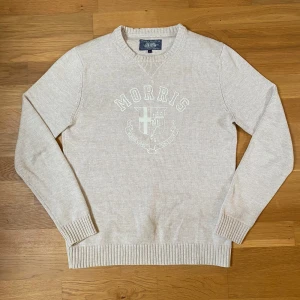 Beige stickad tröja från Morris - Snygg beige stickad tröja från Morris med broderad logga och emblem framtill. Tröjan har rund hals, ribbade muddar och är långärmad. Perfekt för dig som gillar klassisk och stilren look.