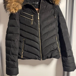 Svart pufferjacka från Hollies med fuskpäls - Snygg svart pufferjacka från Hollies med guldig dragkedja och flera fickor. Jackan har en stor huva med fluffig fuskpäls och quiltad design. Perfekt för kalla dagar när du vill vara både varm och stilren.