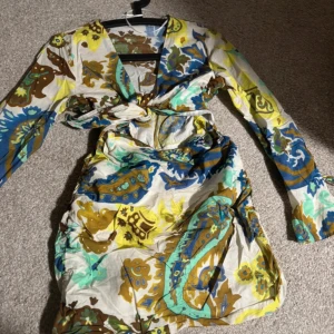 Färgglad omlottklänning med blommigt mönster - Säljer en kort omlottklänning med långa ärmar och v-ringning. Klänningen har ett färgstarkt blommigt mönster i blått, gult, brunt och grönt på vit botten. Knytning i midjan för en snygg passform. Perfekt för dig som gillar unika och färgglada plagg!