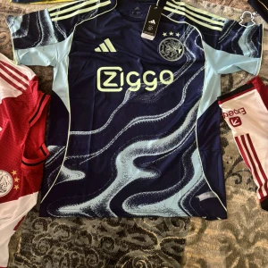 Ajax blå matchtröja Adidas - Ajax fotbollströja från Adidas i mörkblå och ljusblå färg med vågigt mönster. Kortärmad modell med klubbmärke och Ziggo-logga på bröstet. Snygga ljusblå detaljer på axlar och ärmar, tillverkad i lätt polyester som andas. Perfekt för dig som vill sticka ut på planen.