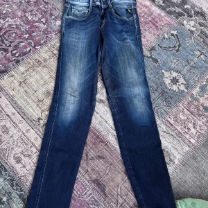 Replay Anbass jeans blå, stl 29/32 - Säljer ett par snygga Replay Anbass jeans i mörkblå tvätt med slitningar och klassiska bakfickor. Modellen har smal passform och raka ben. Jeansen är i stretchigt denim och har detaljerade sömmar vid fickorna. Perfekta för dig som gillar en modern och stilren look.