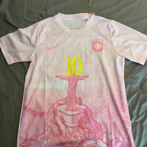 Nike Brasil Neymar JR rosa fotbollströja - Snygg Brasilien fotbollströja från Nike i ljusrosa med gult nummer 10 och Neymar JR på ryggen. Framsidan har en grafisk illustration av Kristusstatyn i Rio och CBF-emblem. Tillverkad i lätt, ventilerande polyester med korta ärmar och rund hals.
