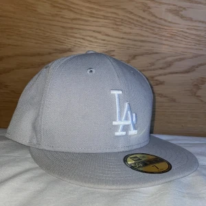 Grå New Era 59FIFTY LA keps - Snygg grå New Era 59FIFTY keps med LA-logga broderad framtill i vitt. Klassisk flat peak och strukturerad passform. Tillverkad i 100% polyester och perfekt för dig som gillar streetwear och sportig stil. Använd en gång