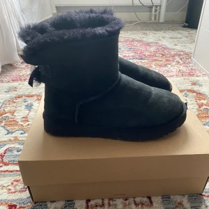 UGG’s - Svarta UGG’s med söta rosetter där bak. Använda max 5 ggr. Supersköna och varma, dom är även inpregnerade så dom är vattentäta. Box medföljer. Nypris är 2650, mitt pris går att diskutera.💗🎀