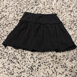 Svart volangkjol med snörning - En svart kortkjol med volang och snörning i midjan. Kjolen har en avslappnad passform och är gjord i mjuk bomull, perfekt för en chill och trendig look. Elastisk midja gör den bekväm och enkel att bära.