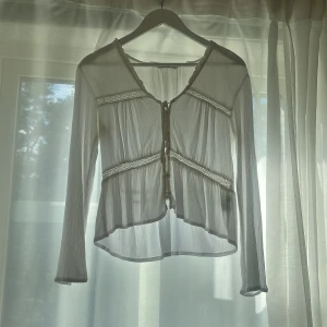 Vit blus med knyt och spetsdetaljer - Superfin vit blus i tunn, plisserad bomull med långa ärmar och v-ringning. Blusen har knyt framtill och dekorativa spetsband.