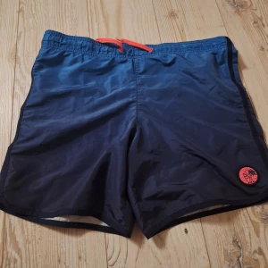 Mörkblå badshorts från H&M - Snygga mörkblå badshorts från H&M med röd snörning i midjan och ett coolt orange märke med palmmotiv på benet. Shortsen har en loose passform och är gjorda i lätt syntetmaterial som torkar snabbt. Perfekta för strandhäng och pool.