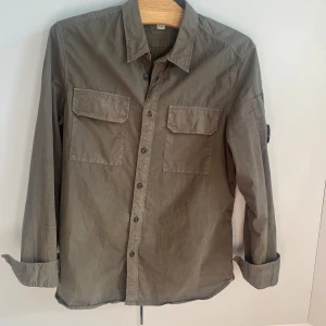 Olivgrön overshirt från C.P. Company - Snygg olivgrön overshirt från C.P. Company med klassisk krage, två bröstfickor med lock och knappar framtill. Är strl L men sitter som en M