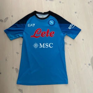 Napoli blå fotbollströja EA7 - Snygg Napoli fotbollströja från EA7 i blått med svarta axlar och vita samt röda tryck. Kortärmad modell med rund hals och diskret mönster i tyget. Perfekt för dig som vill representera Napoli på match eller träning.