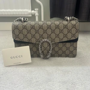 Gucci Dionysus axelväska monogram - Säljer en Gucci Dionysus axelväska med klassiskt GG-monogram i beige och brunt. Väskan har en silverfärgad kedja och ikoniskt spänne med tigrar framtill. Tillverkad i canvas med skinn-detaljer och snyggt mönster över hela väskan. Perfekt för dig som vill sticka ut.