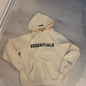 Essentials hoodie i beige med tryck - Beige hoodie från Essentials Fear of God med svart tryck på bröstet och ärmen. Klassisk känguruficka, ribbade muddar och huva. Oversized fit som ger en chill vibe, perfekt till jeans eller sweatpants.